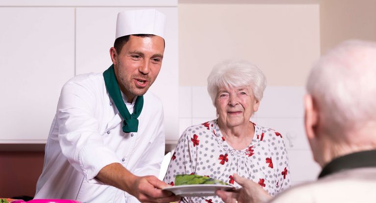 Koch mit Mütze und grünem Halstuch serviert einem älteren Paar Essen