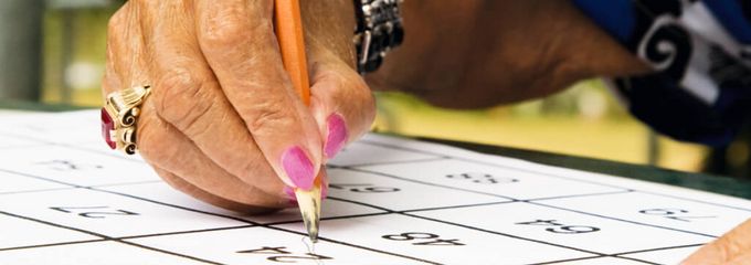 Ältere Frau mit rosa Fingernägeln füllt Bingo-Papier mit einem Bleistift aus