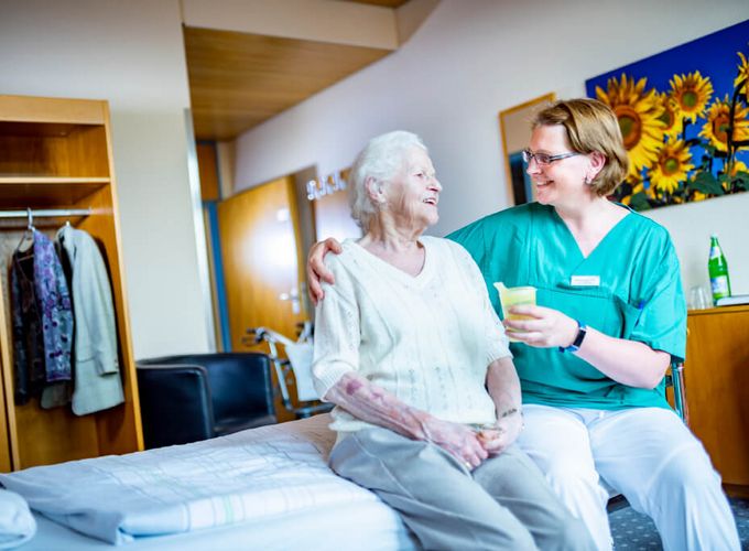 Aktivierende Pflege Pflegekraft reicht Seniorin einen Becher mit Saft, während Sie auf dem Bett sitzen